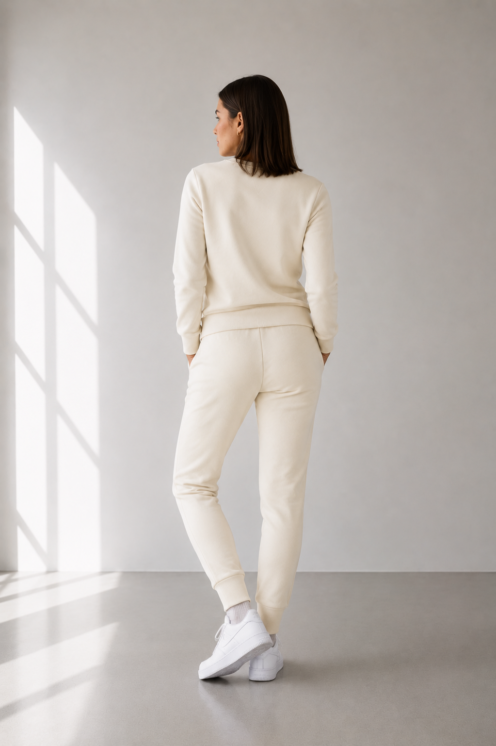Person wearing a cream-colored outfit standing in a minimalistic room with a large window.