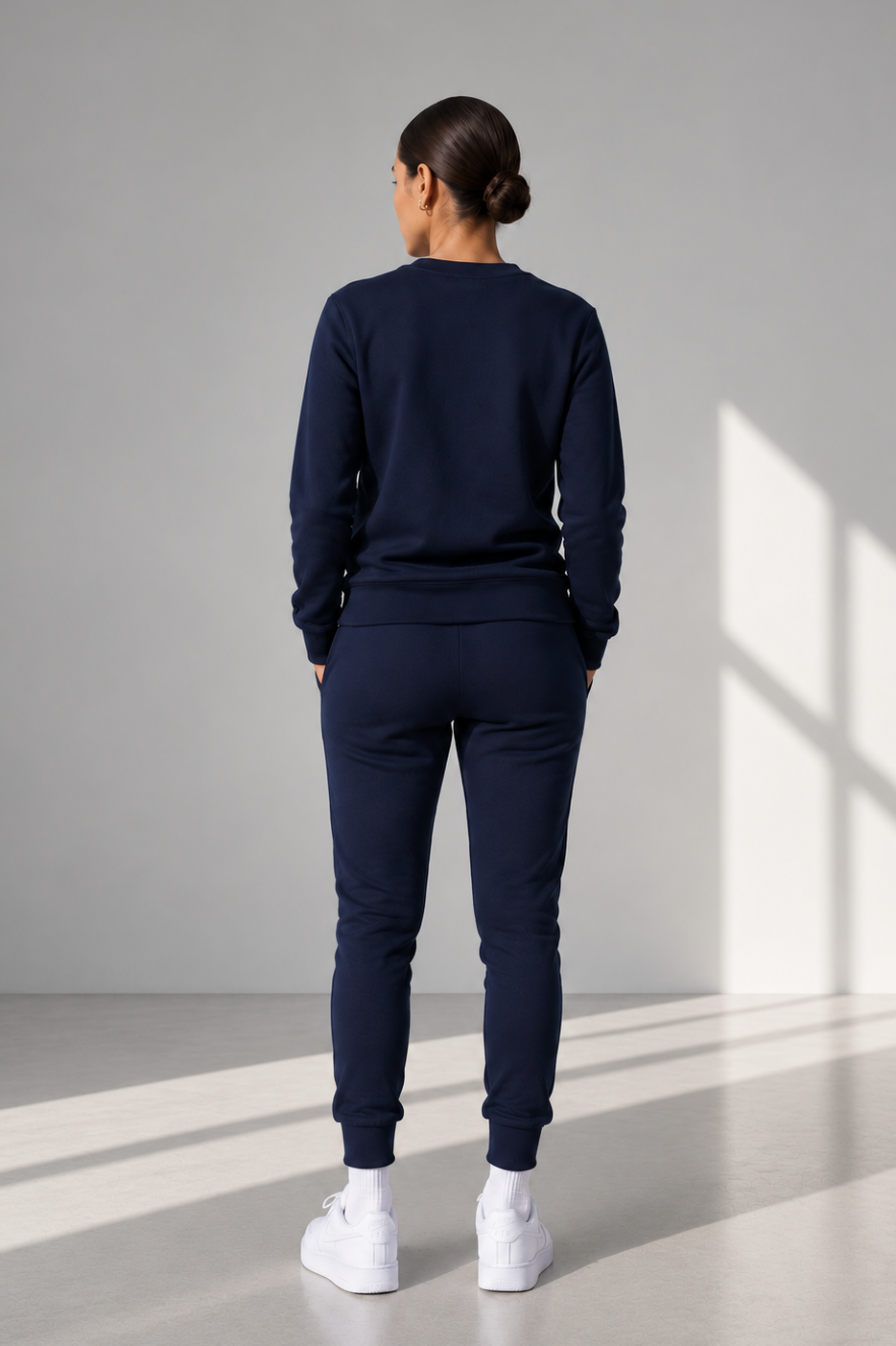 A woman viewed from behind wearing a matching navy blue sweatshirt and jogger pants set, paired with white sneakers, standing in a minimalist gray studio with window-light shadows.