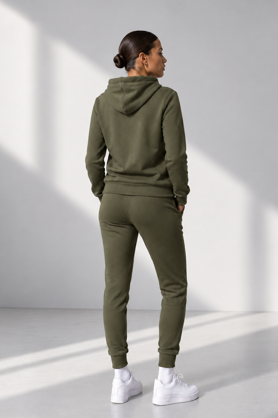 A woman wearing an olive green hooded sweatshirt and matching jogger pants, shown from the back in a minimalist studio setting with soft diagonal shadows.