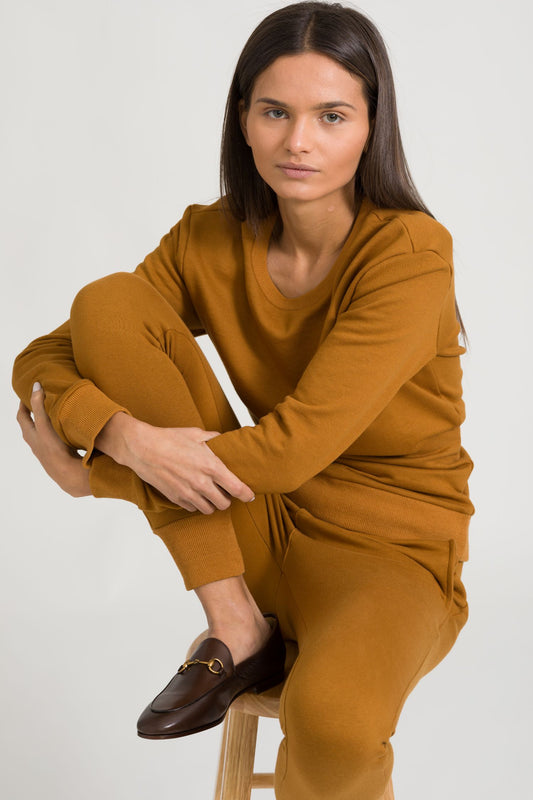 Woman wearing a mustard yellow outfit sitting on a stool against a white background