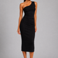 A woman modeling a black one-shoulder midi dress with a peplum waist, paired with white heeled sandals, against a solid gray studio background.