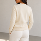 A woman shown from the back wearing a cream-colored crewneck sweatshirt and matching cream sweatpants in a minimalist studio setting.
