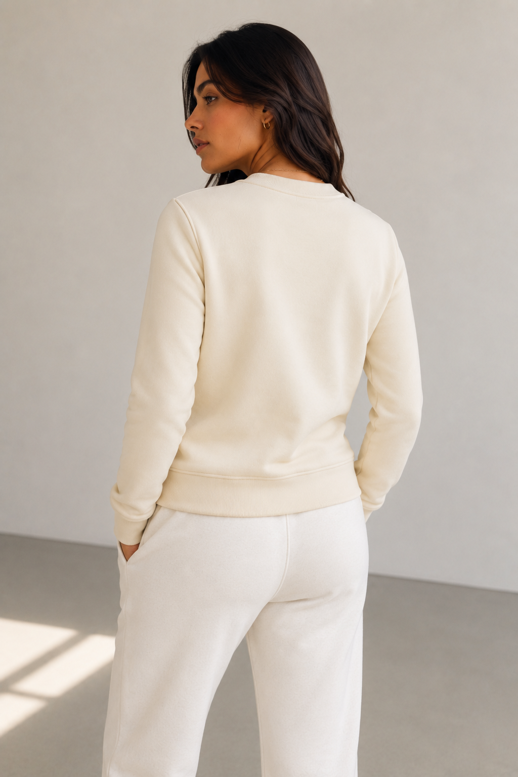 A woman shown from the back wearing a cream-colored crewneck sweatshirt and matching cream sweatpants in a minimalist studio setting.