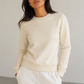 A woman wearing a cream-colored crewneck sweatshirt and matching cream sweatpants, standing against a neutral light gray wall.