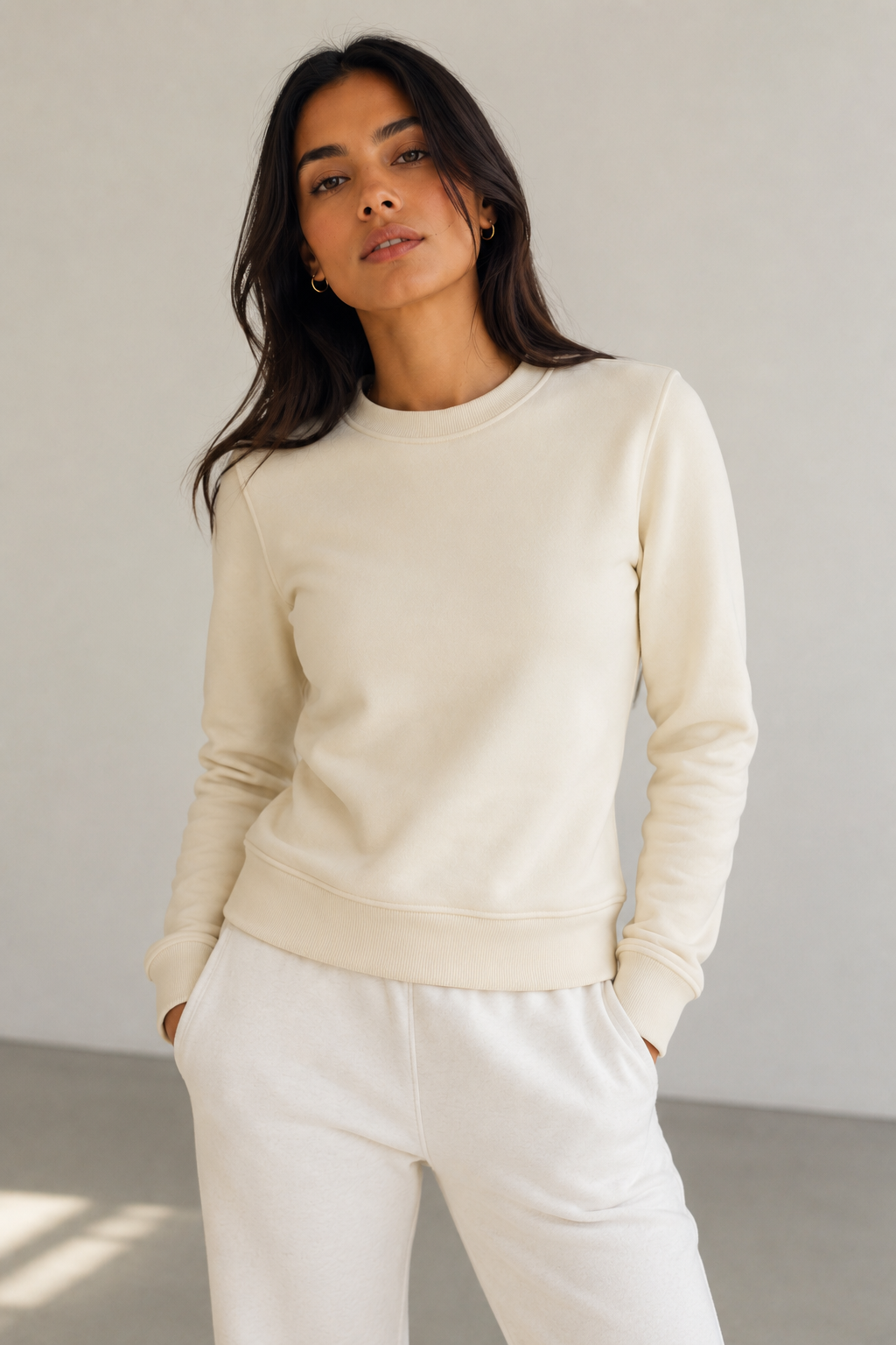 A woman wearing a cream-colored crewneck sweatshirt and matching cream sweatpants, standing against a neutral light gray wall.