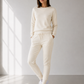 Woman wearing a cream-colored outfit standing in a bright room with large windows.
