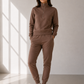 A woman modeling a matching brown lounge set consisting of a half-zip sweatshirt and jogger pants, paired with white sneakers, in a minimalist room with window shadows.