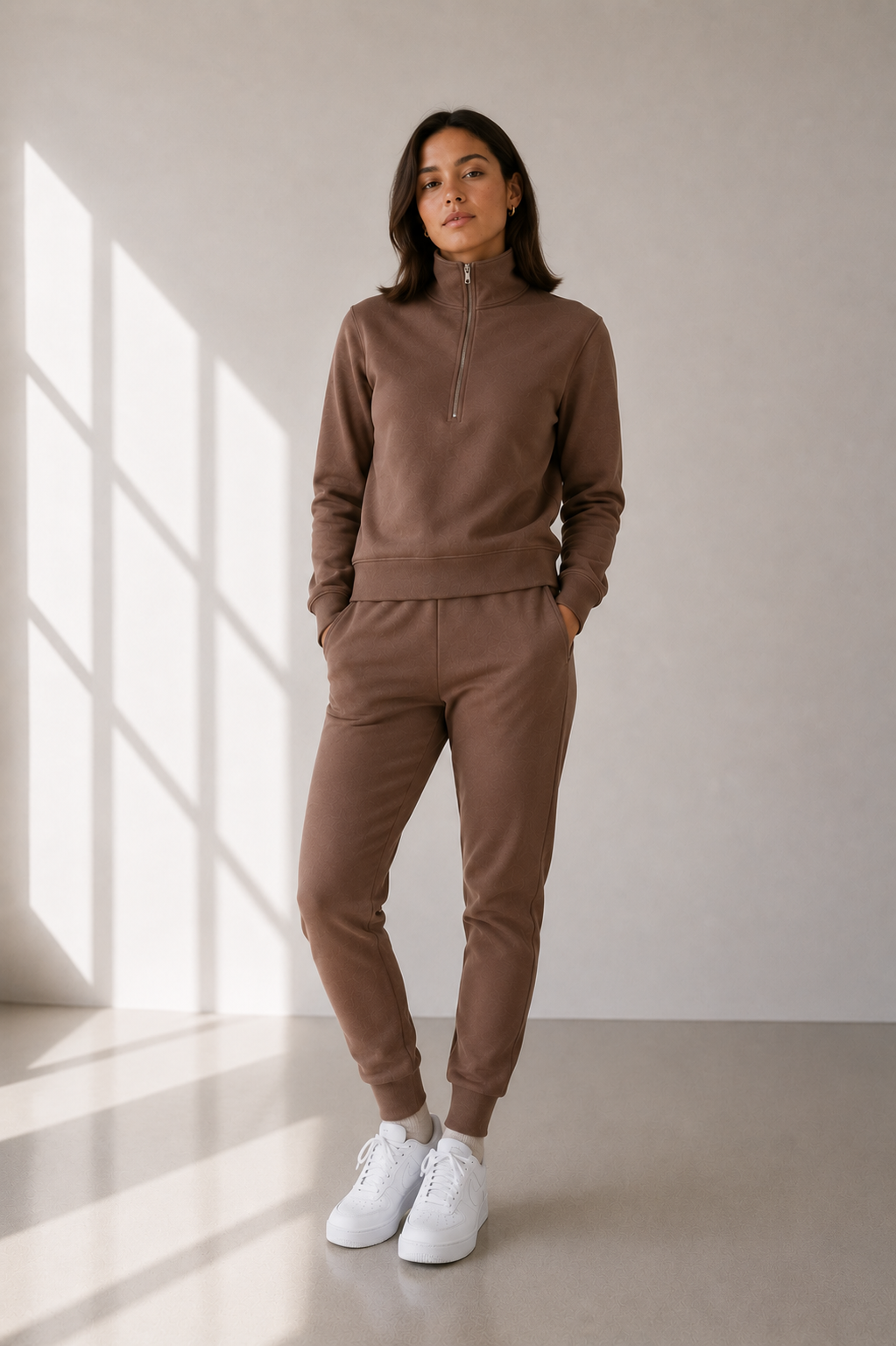 A woman modeling a matching brown lounge set consisting of a half-zip sweatshirt and jogger pants, paired with white sneakers, in a minimalist room with window shadows.
