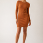 A woman modeling a fitted, long-sleeve mini dress in a warm camel brown color, paired with matching brown strappy high-heeled sandals, against a plain light gray studio background.