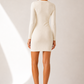A woman stands with her back to the camera wearing a fitted long-sleeve mini dress in a warm off-white/cream color, paired with white sneakers, against a minimalist beige studio backdrop with soft diagonal light rays.