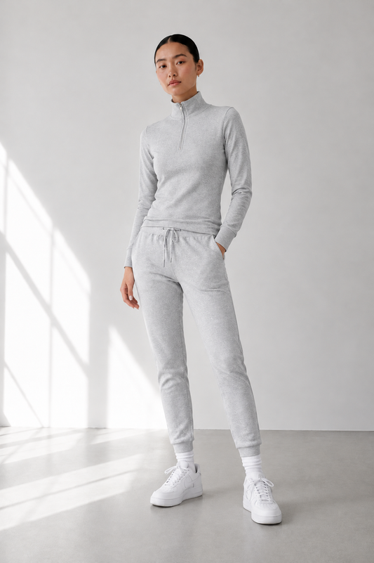 A woman modeling a light gray heathered two-piece tracksuit consisting of a mock neck quarter-zip long-sleeve top and matching jogger pants, paired with white sneakers in a bright, minimalist studio.