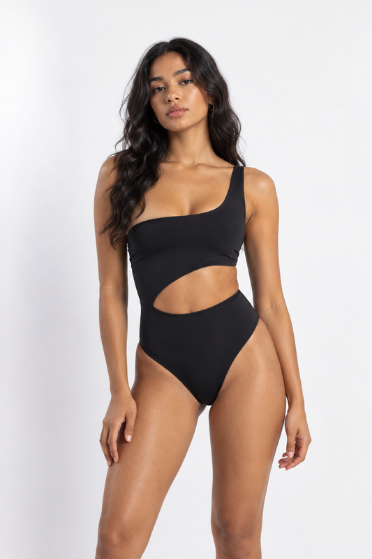 A woman modeling a black one-shoulder cutout one-piece swimsuit against a plain light gray studio background.