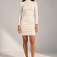 A woman modeling a fitted, long-sleeve cream-colored mock-neck mini dress with a zippered high collar, paired with white sneakers, against a neutral studio backdrop.