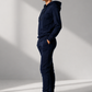 A woman stands in profile wearing a matching navy blue hoodie and jogger sweatpants set with white sneakers, photographed in a minimalist studio with dramatic diagonal lighting.