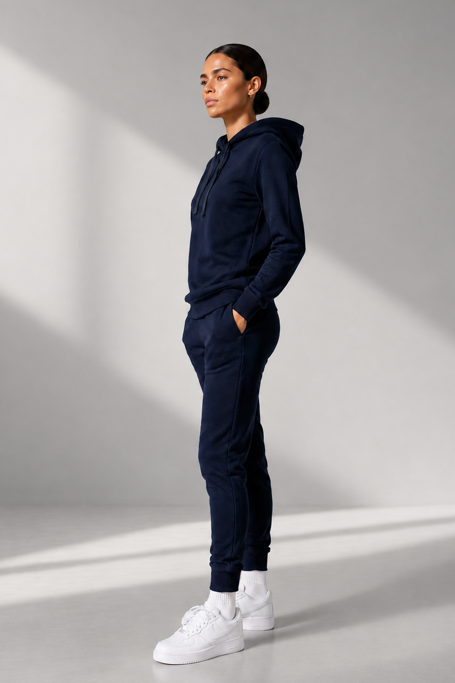 A woman stands in profile wearing a matching navy blue hoodie and jogger sweatpants set with white sneakers, photographed in a minimalist studio with dramatic diagonal lighting.