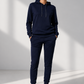 A model wearing a matching navy blue sweatshirt and jogger pants set, paired with white sneakers, standing in a minimalist studio with soft window light shadows.
