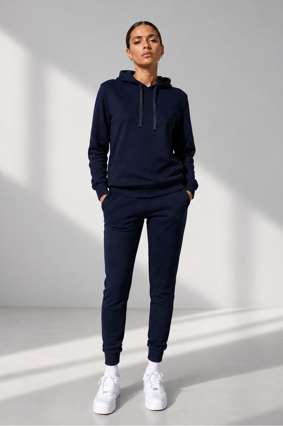 A model wearing a matching navy blue sweatshirt and jogger pants set, paired with white sneakers, standing in a minimalist studio with soft window light shadows.