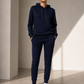 A model stands in a minimalist studio wearing a matching navy blue hoodie and jogger pants set, paired with white sneakers.