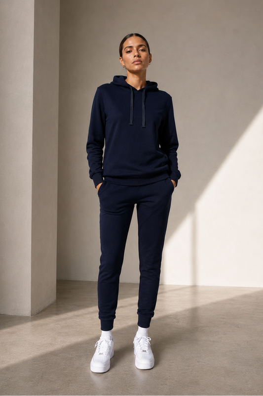 A model stands in a minimalist studio wearing a matching navy blue hoodie and jogger pants set, paired with white sneakers.