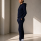 A woman stands in profile wearing a matching navy blue hoodie and jogger sweatpants set with white sneakers, posed in a minimalist studio with warm sunlight and shadows.
