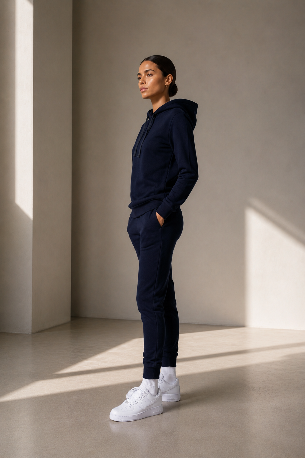 A woman stands in profile wearing a matching navy blue hoodie and jogger sweatpants set with white sneakers, posed in a minimalist studio with warm sunlight and shadows.