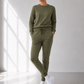 A woman modeling a matching olive green lounge set consisting of a long-sleeve crewneck sweatshirt and drawstring jogger pants, paired with white sneakers in a bright, modern room with window light.