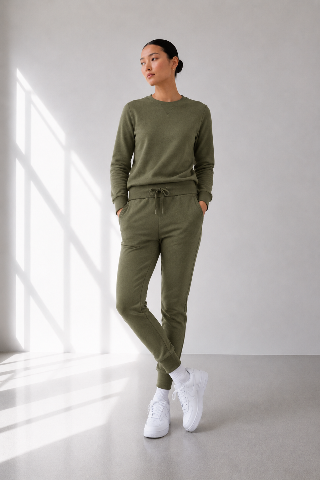 A woman modeling a matching olive green lounge set consisting of a long-sleeve crewneck sweatshirt and drawstring jogger pants, paired with white sneakers in a bright, modern room with window light.
