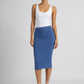 A model stands against a light gray studio background wearing a white sleeveless top and a fitted blue midi skirt, paired with clear high-heeled sandals.