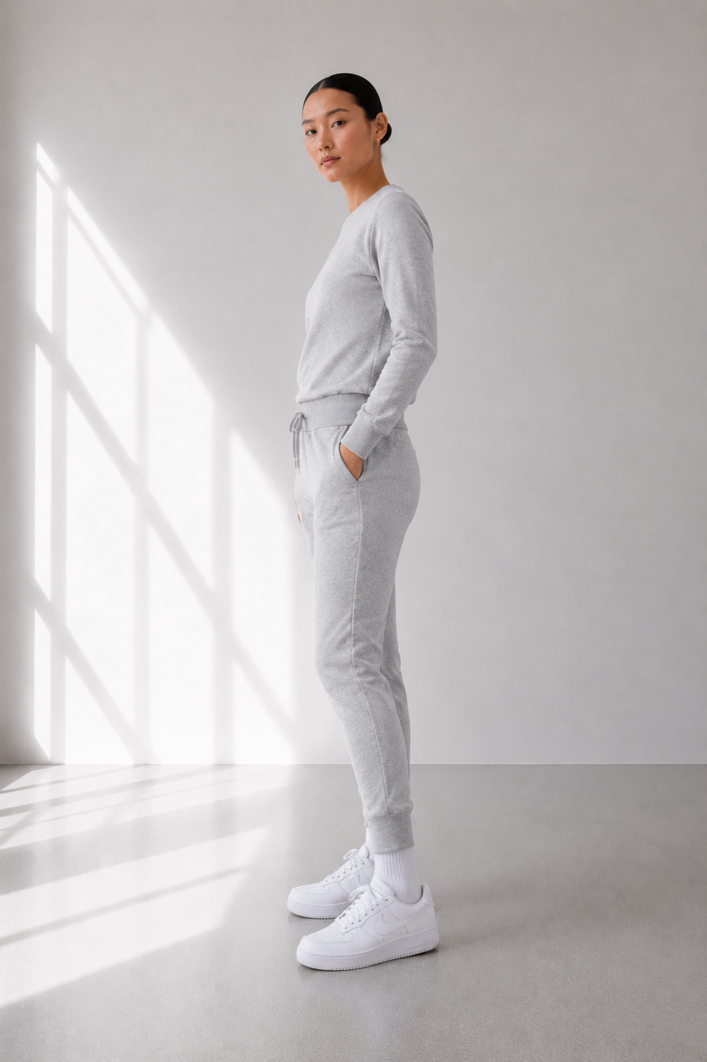 Person wearing a light gray outfit standing in a minimalistic room with a large window.