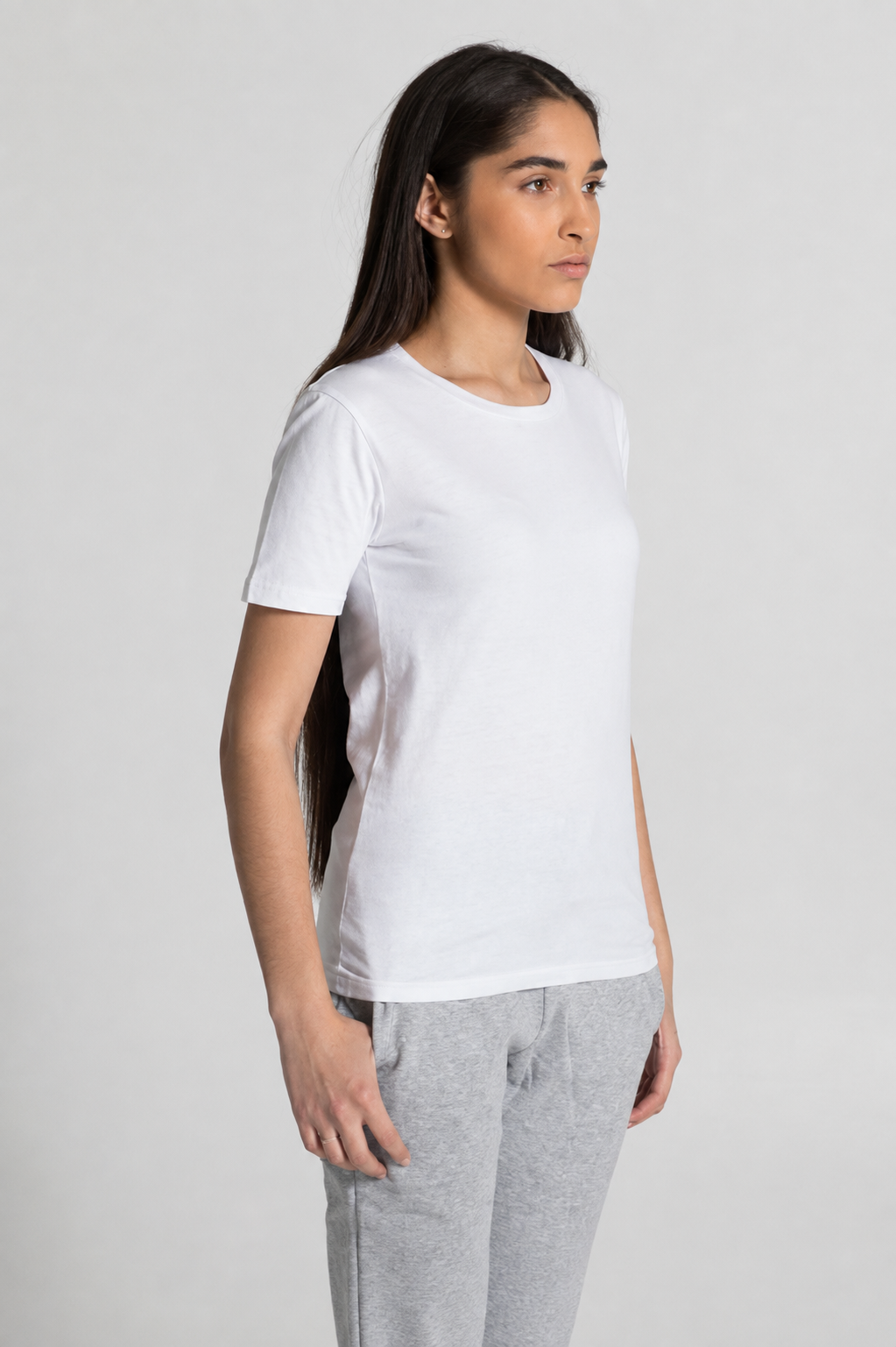 A woman wearing a plain white short-sleeve crewneck t-shirt and light gray sweatpants, standing against a light gray studio background.