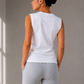 A woman shown from behind wearing a white sleeveless t-shirt and light gray heathered leggings in a minimalist gray room with window shadows.